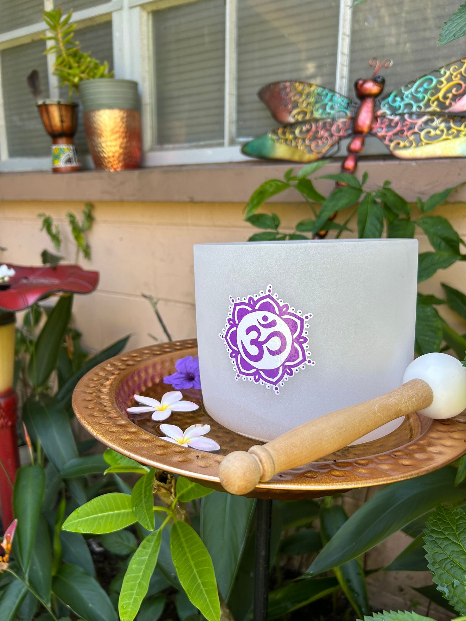 Sacred frosted singing bowl with purple Om symbol in lotus mandala design, surrounded by floating plumeria flowers and lush greenery